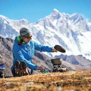Eine Frau kocht mit ihrem Gaskocher auf einer Anhöhe, im Hintergrund Berge.