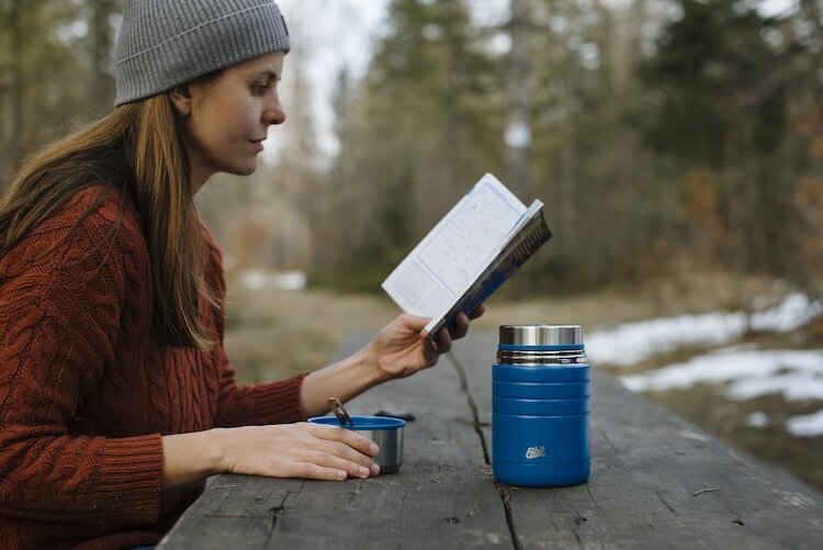 Frau liest im Wald in einem Buch, vor ihr steht ein blauer Esbit Thermobecher 
