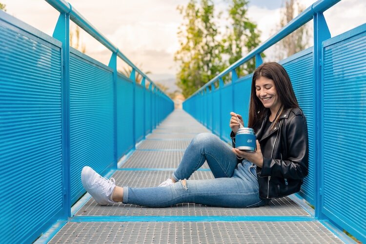 Junge Frau sitzt auf einer Brücke und isst aus einem Esbit Thermobecher 